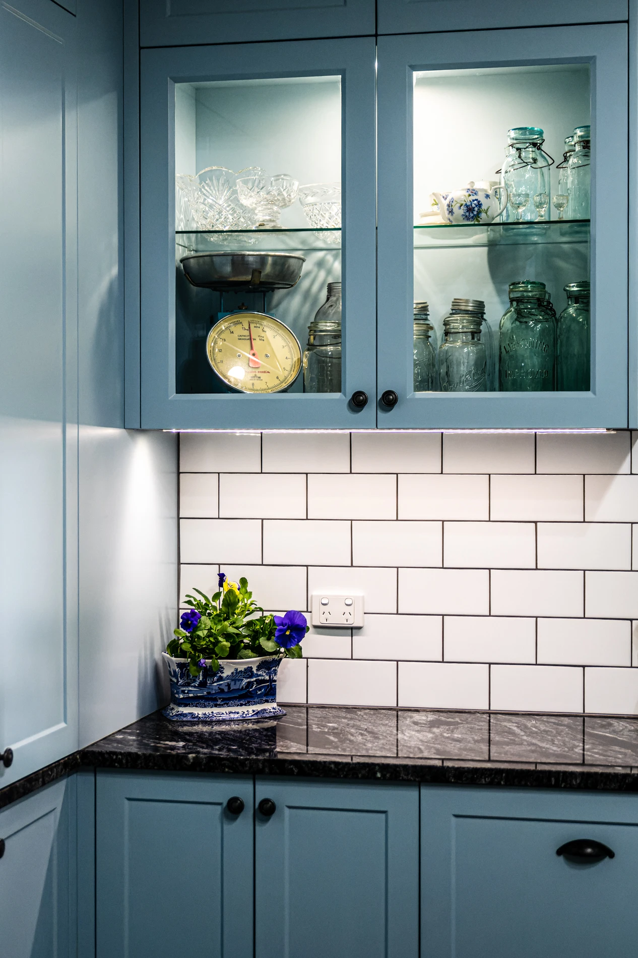 Blue Cabinet Corner With Glassware, Decorative Scale; Planter With Flowers On Marble Countertop.