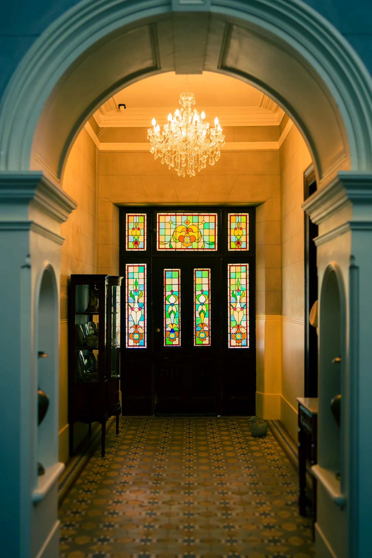 Elegant Hallway With Chandelier, Patterned Tiles, Arched Entrance, And Stained Glass Windows.