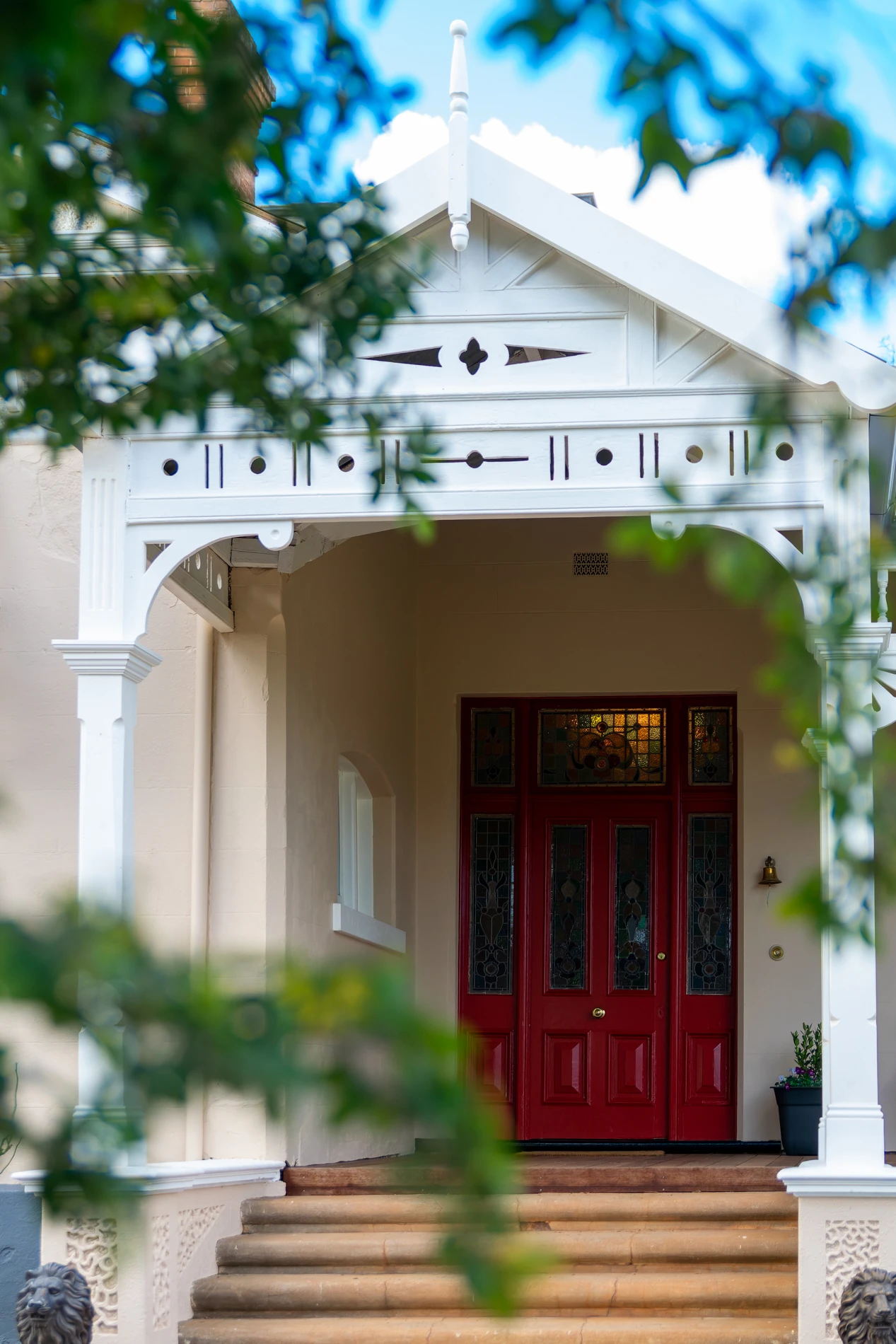 House Entrance With Veranda, Red Door, Ornate Glass Panels, Columns, Steps, And Surrounding Foliage.