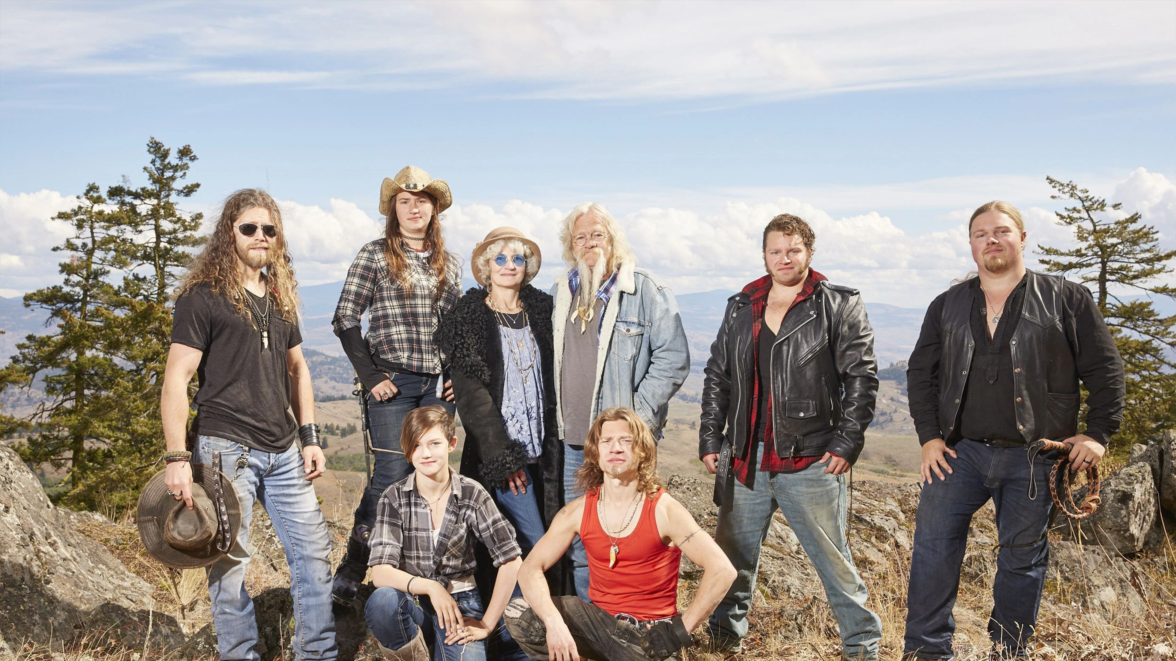 Group On Rocky Hill With Mountainous Backdrop, Wearing Casual Rugged Attire, Clear Cloudy Sky.
