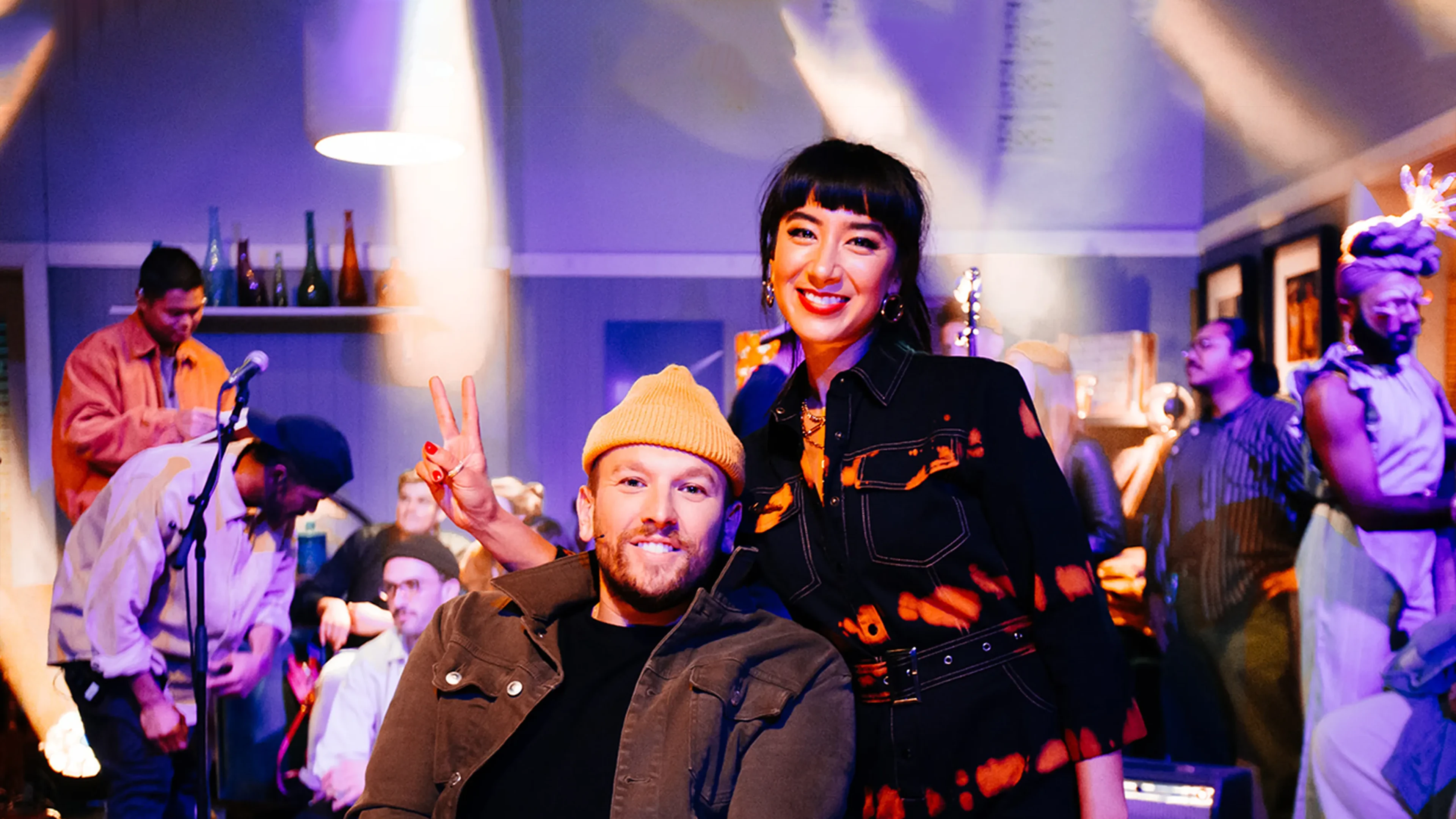 Two smiling people in front at a lively, dim social event with stage and microphones.