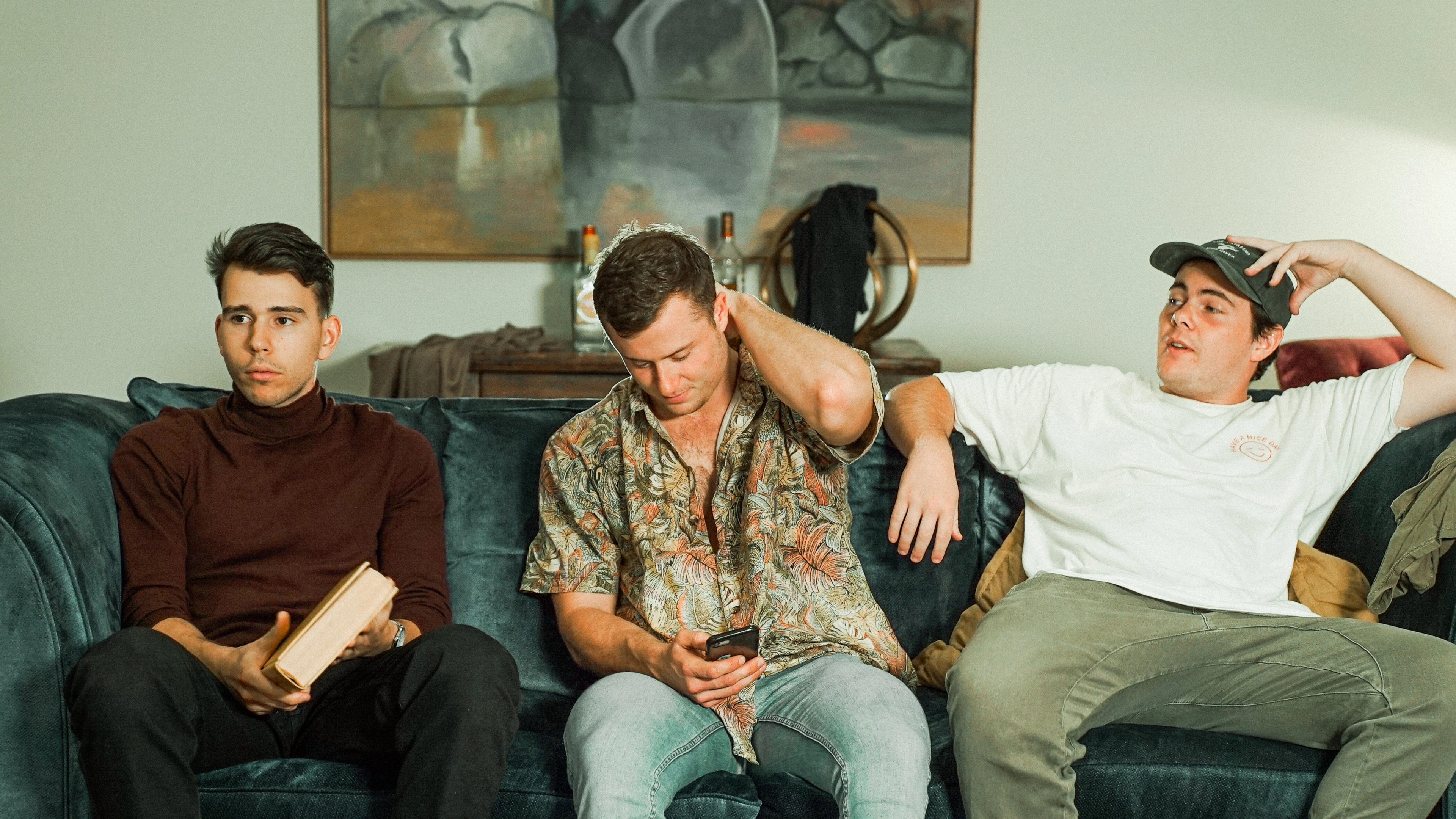 Three men on a blue couch in a living room, with a book, phone, cap, painting, and bottles.