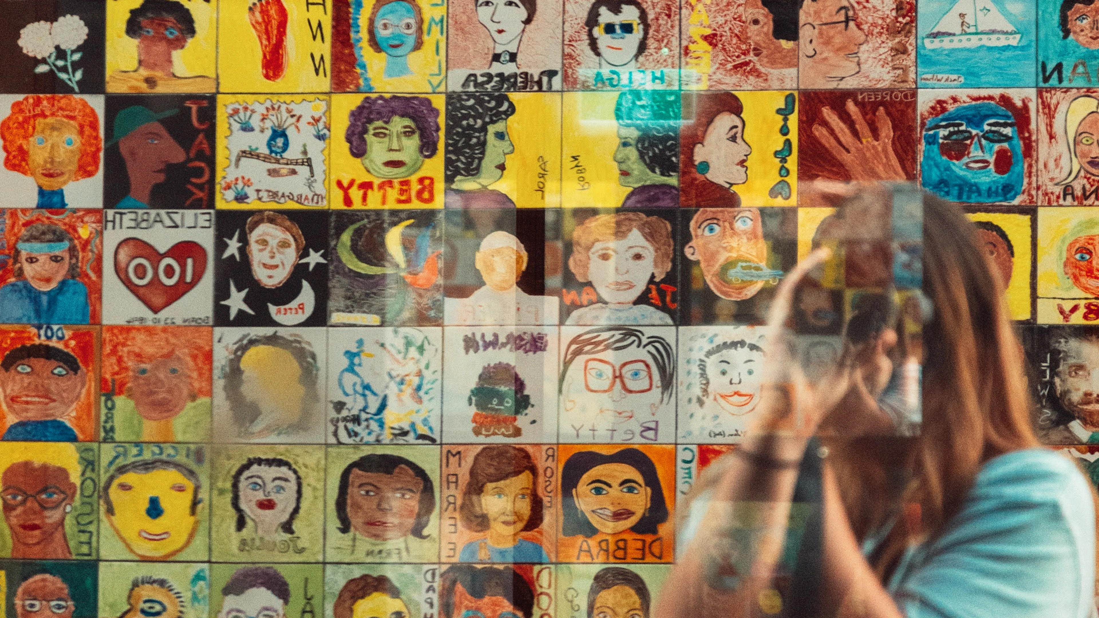 Person reflected in glass, standing before a vibrant mural of diverse painted portraits.