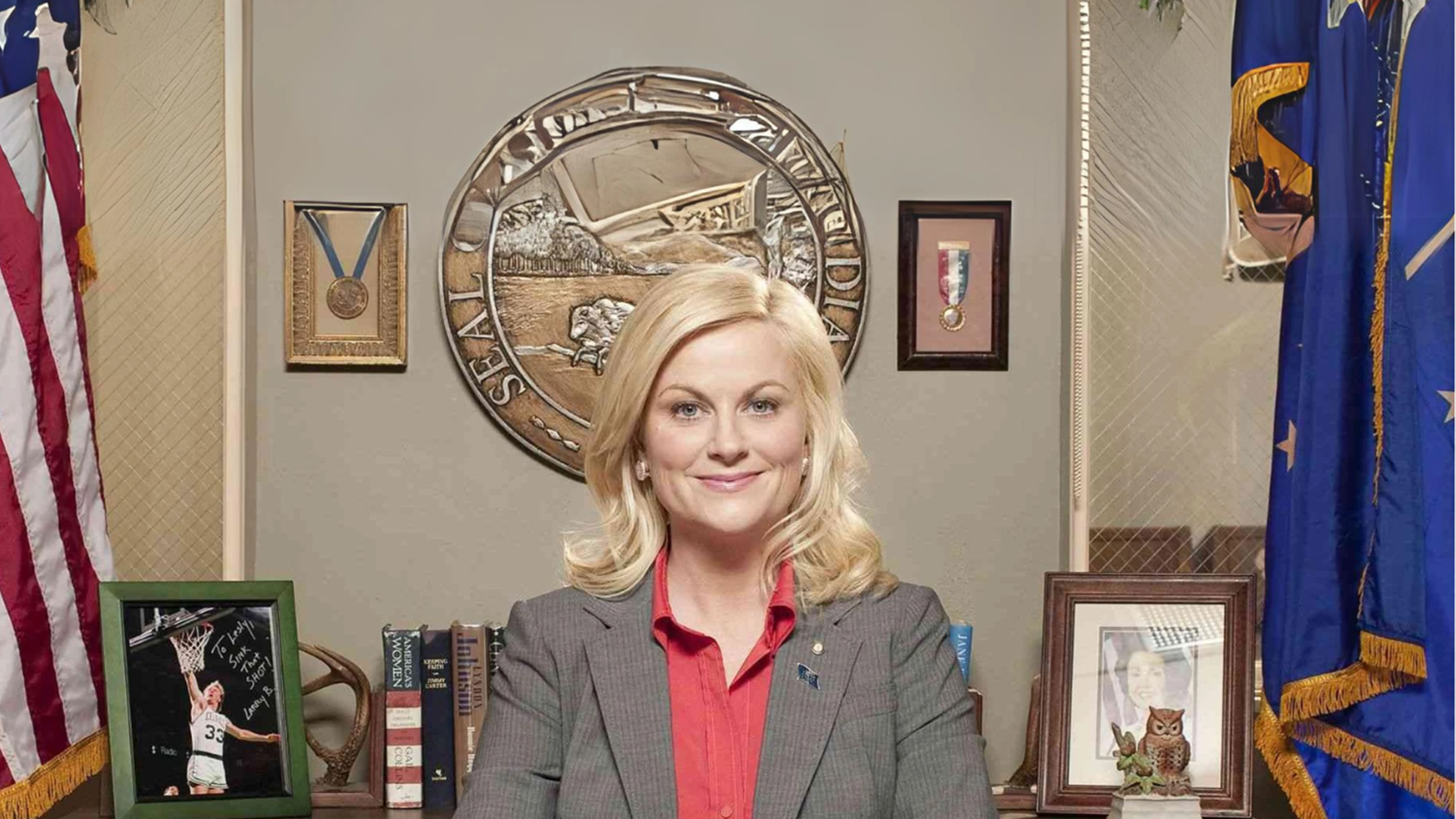 Blonde person at office desk, large seal, framed medals, certificates, flags, owl figurine visible.