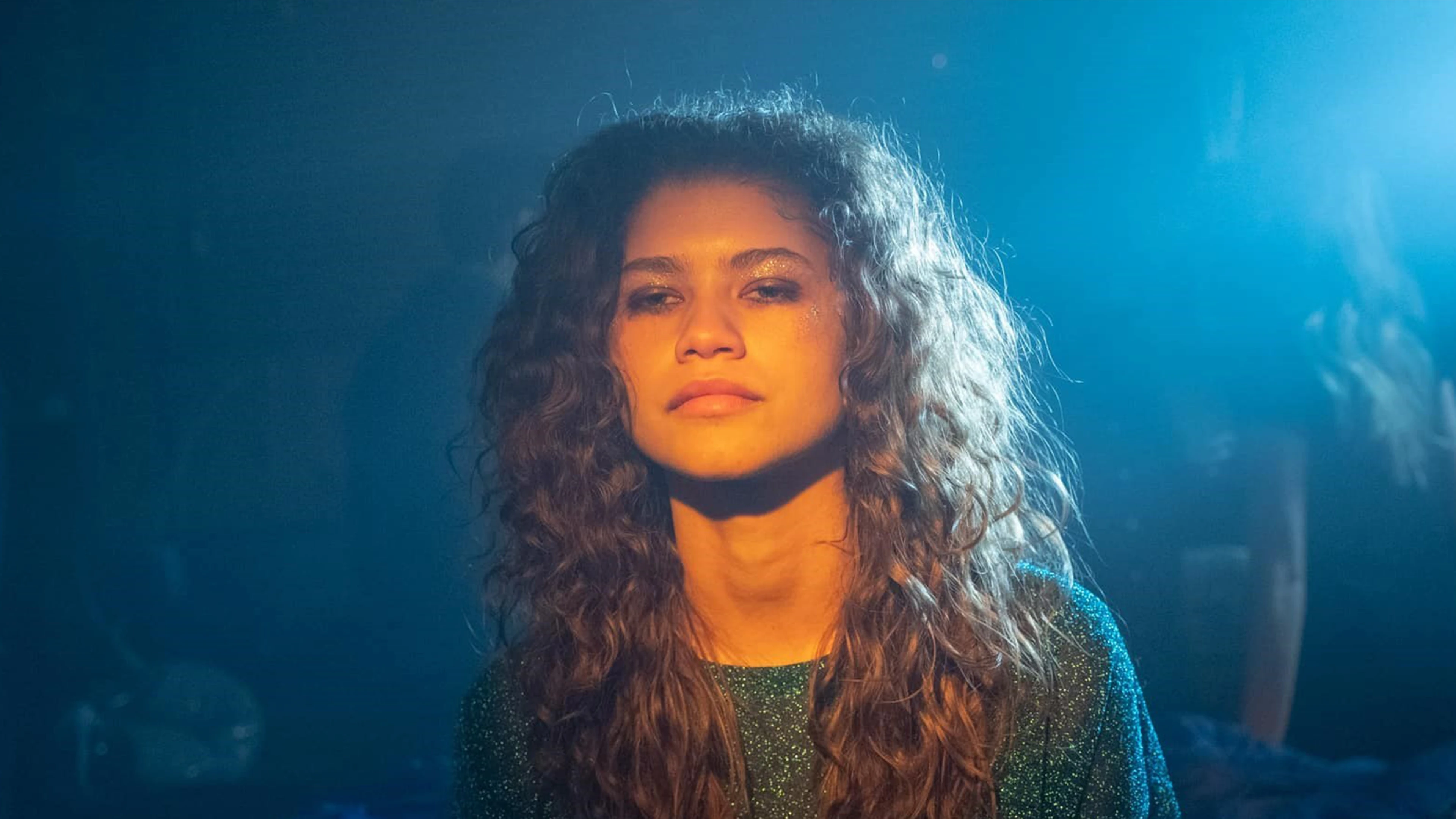 About Euphoria: Which Character Sucks The Most? 9 Person With Curly Hair In Dim Lighting, Blue Background, Wearing Dark Top, Neutral Expression.