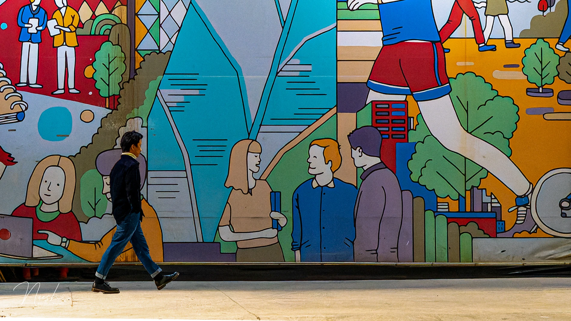 Man Walking By A Colourful Mural With Stylised Illustrations Of People, Trees, And Patterns.