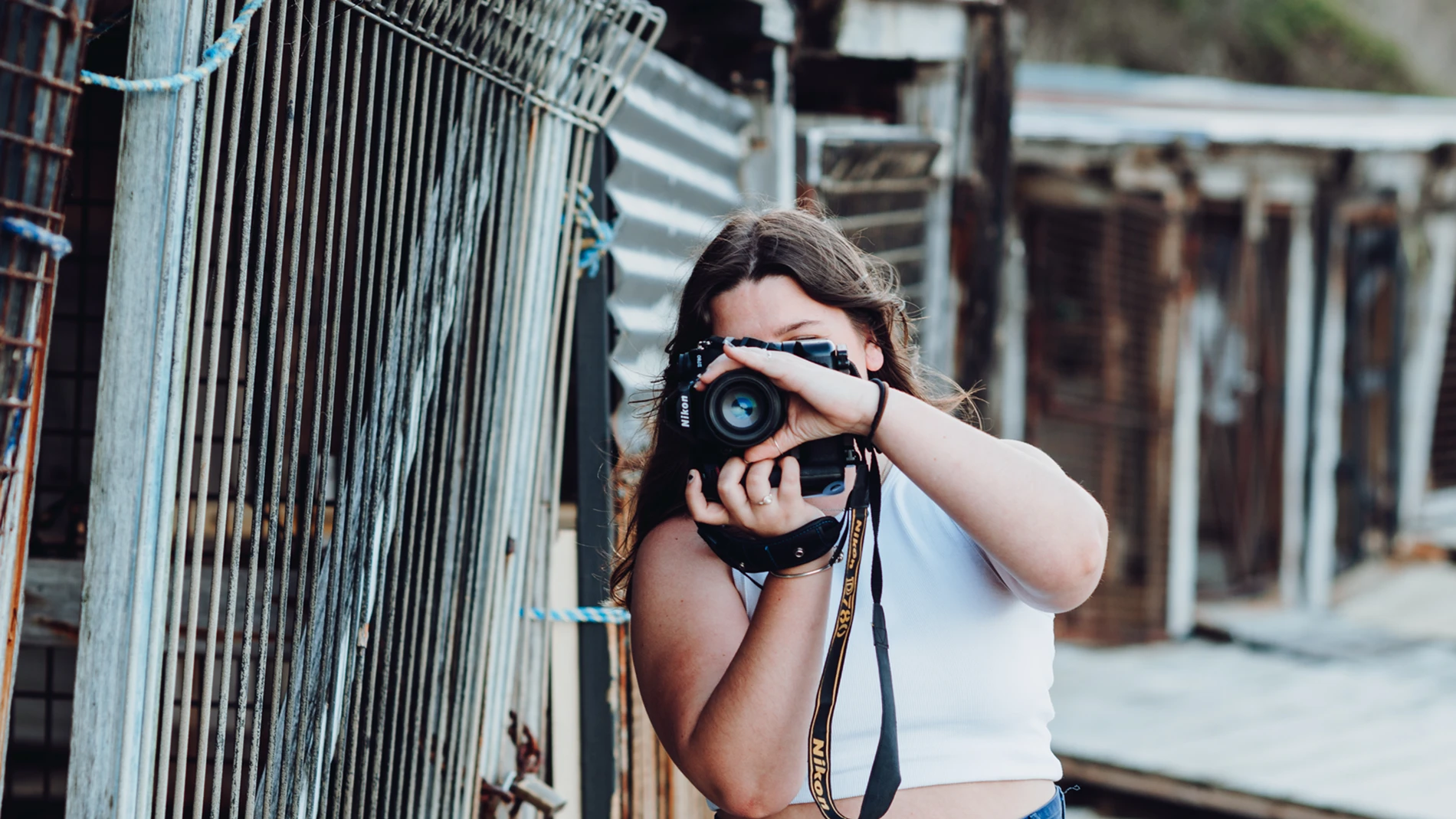 Person With Long Hair Takes Photo Near Metal Fence And Wooden Structures Outdoors.