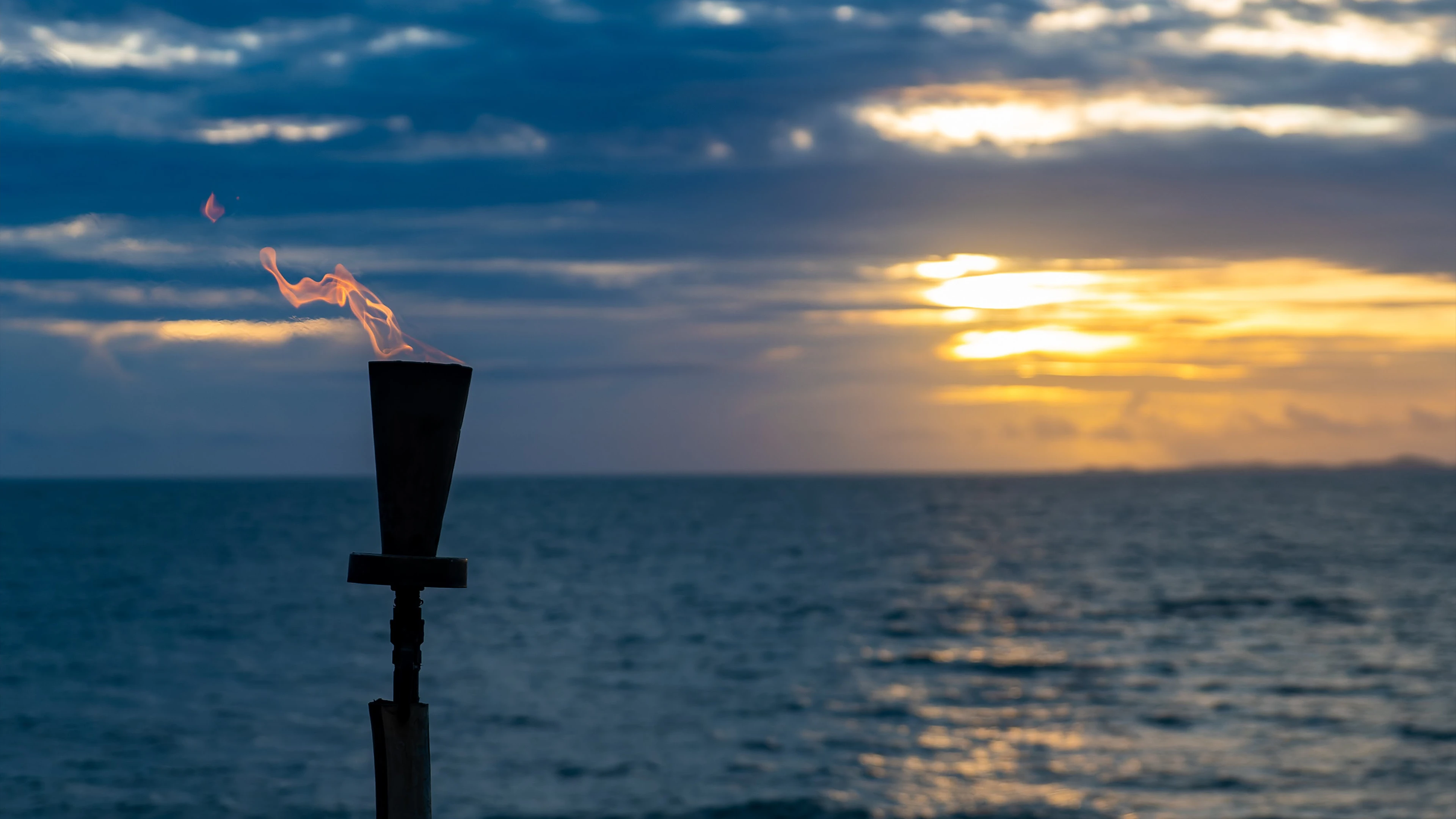 Torch With Flame By An Ocean Sunset, Sky Filled With Clouds And Warm Colours.