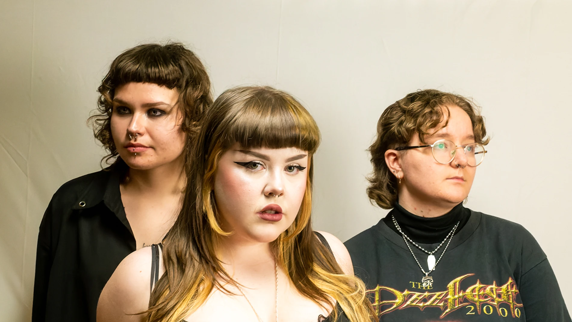 Three Individuals With Unique Hairstyles And Outfits Stand Together Against A Plain Background.