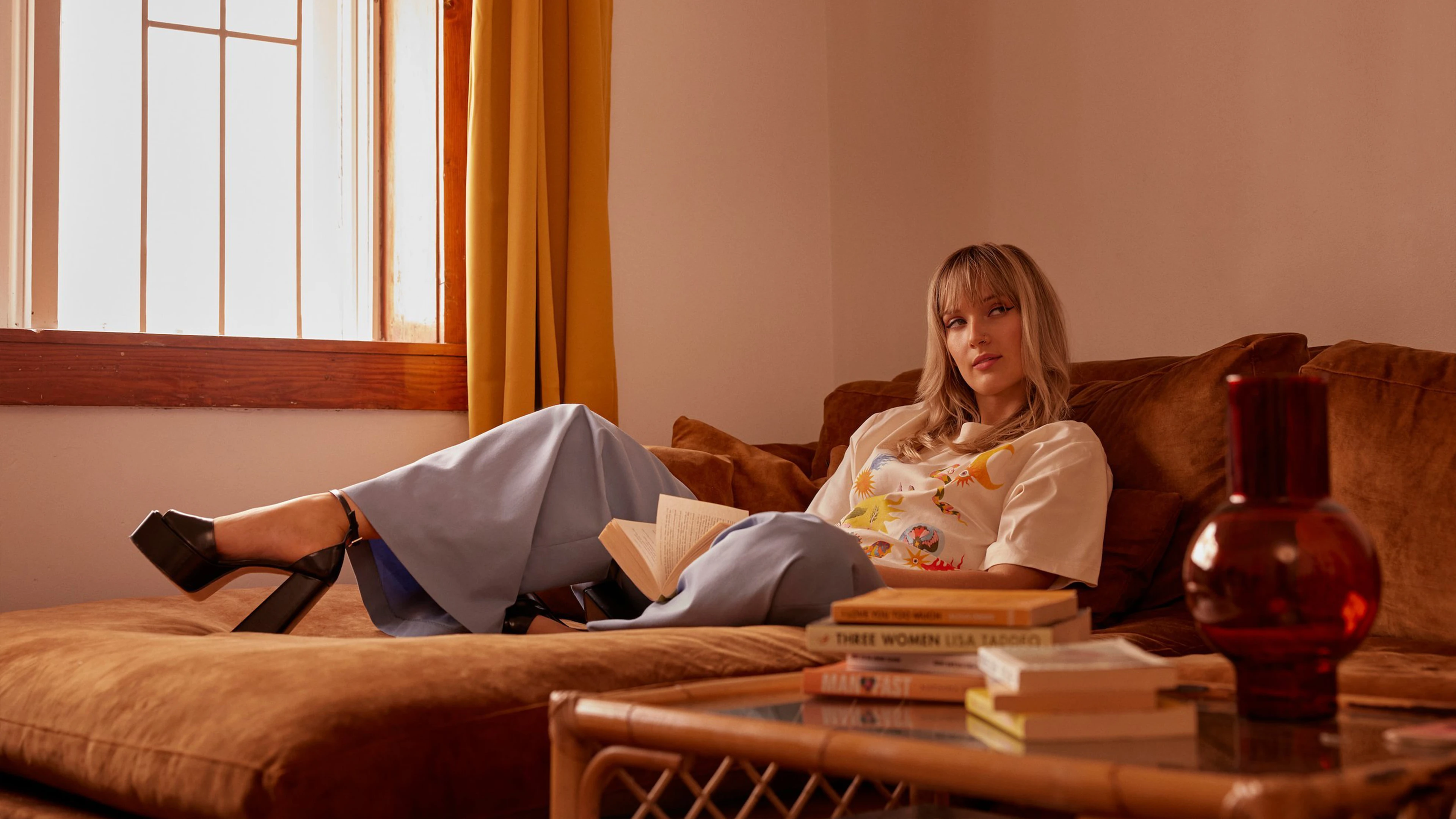 Person On Brown Sofa With Book, Wearing Blue Trousers, Patterned Shirt; Window And Table Visible.