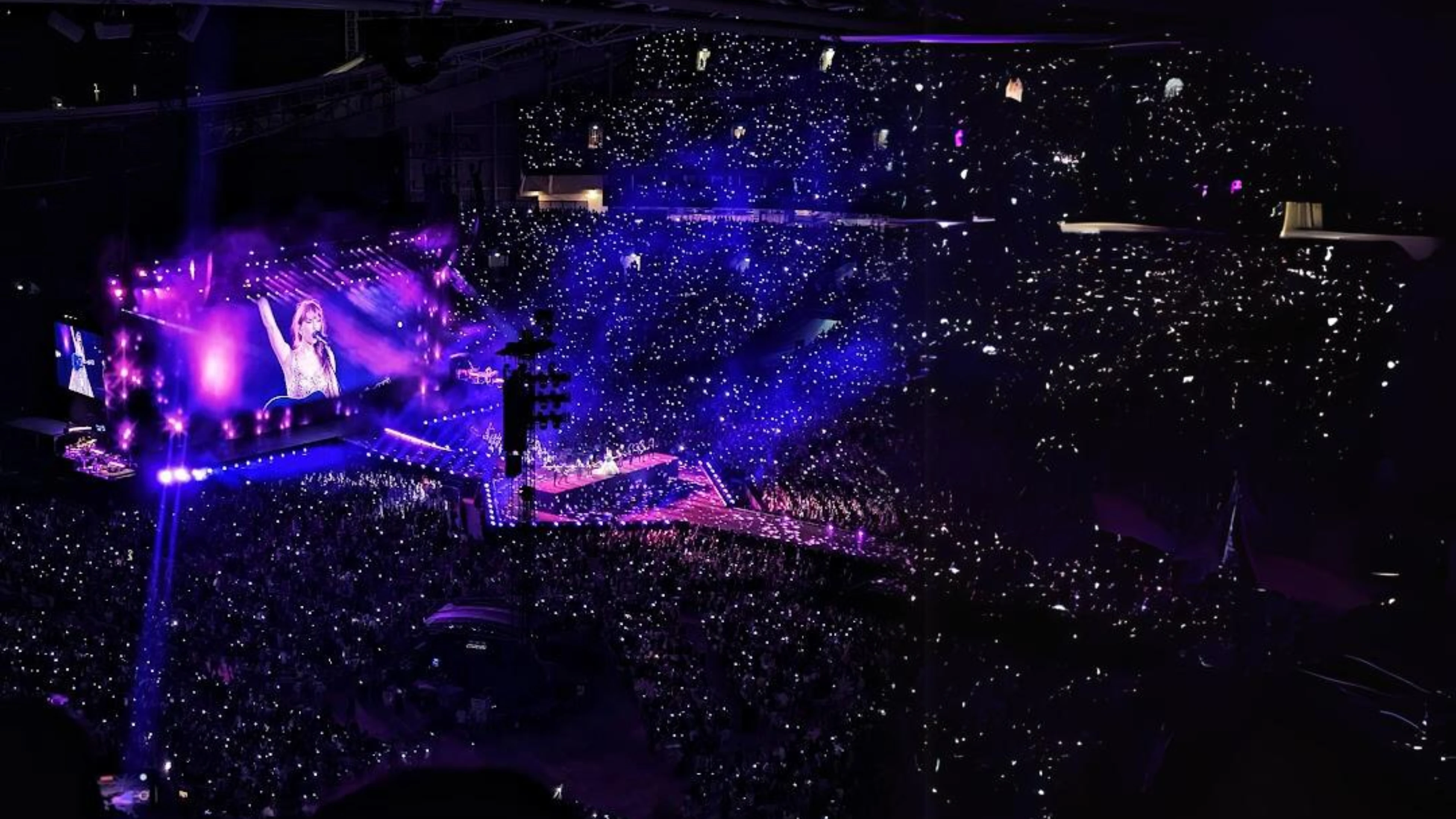 Large concert with vibrant light display, performer on screen, and illuminated audience in stadium.