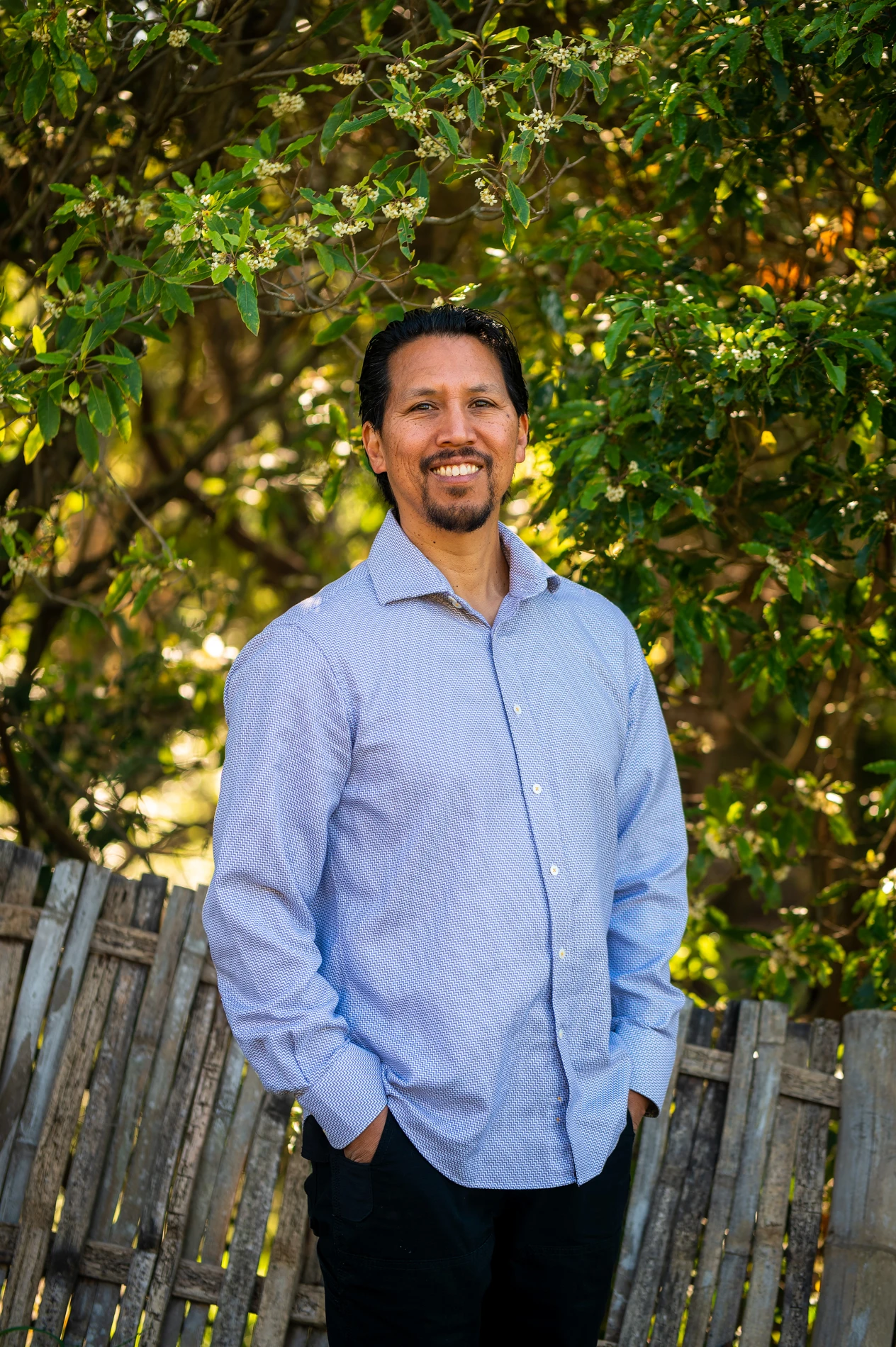 Man Smiling Outdoors, In Light Blue Shirt, Black Trousers, With Green Foliage And Wooden Fence Behind.