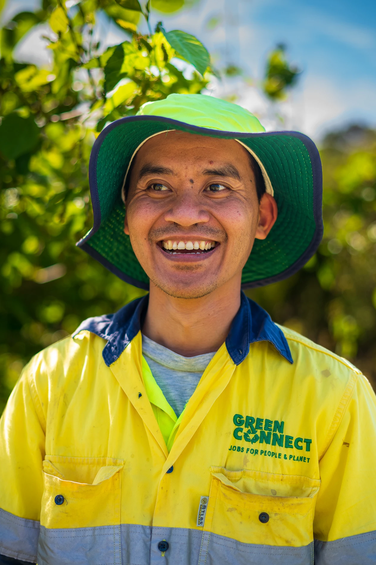 Person In Yellow-Blue Work Shirt And Green Hat Smiling, With Greenery And Clear Sky Background.