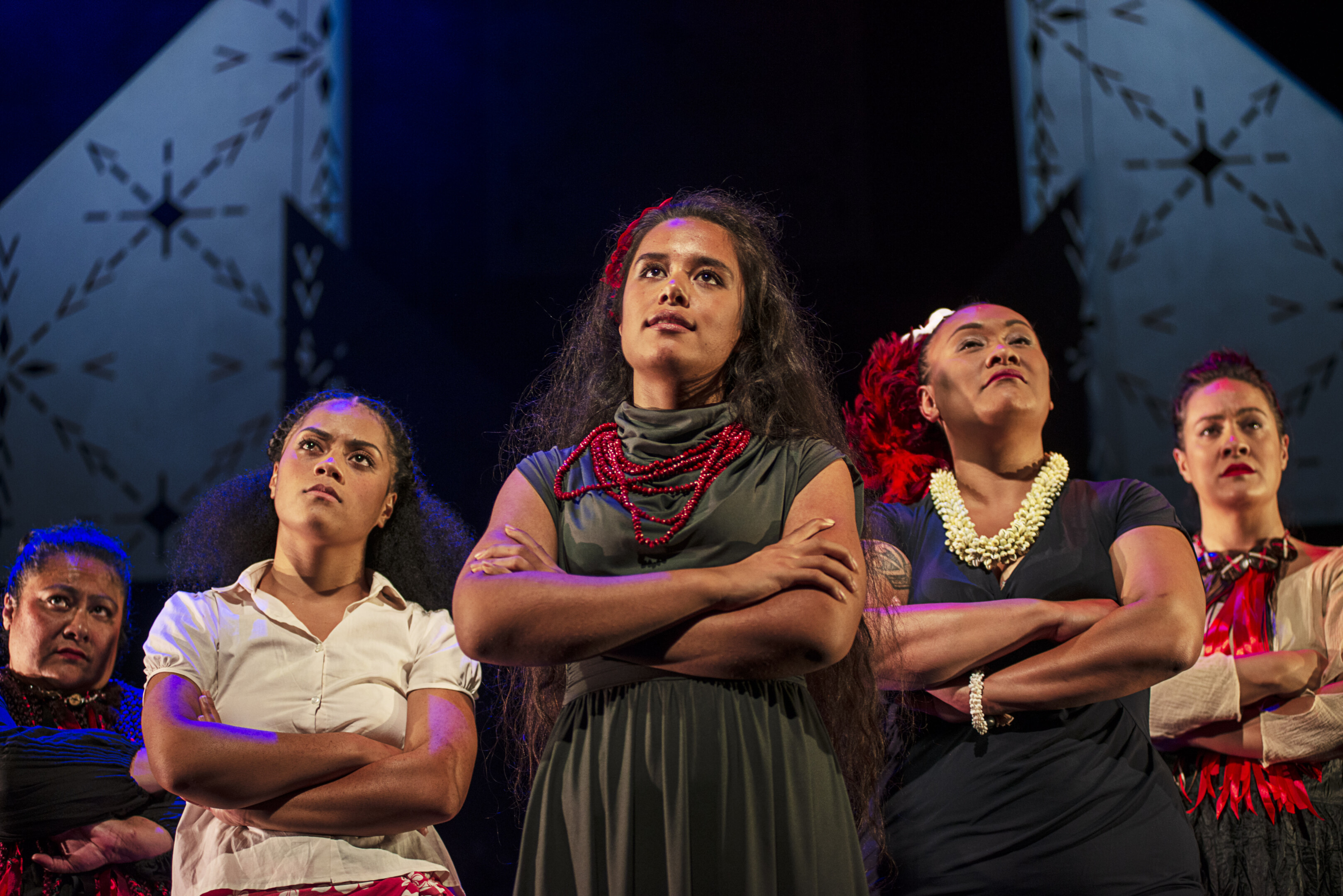 Group on stage in traditional attire, arms crossed, dramatic lighting, patterned backdrop.