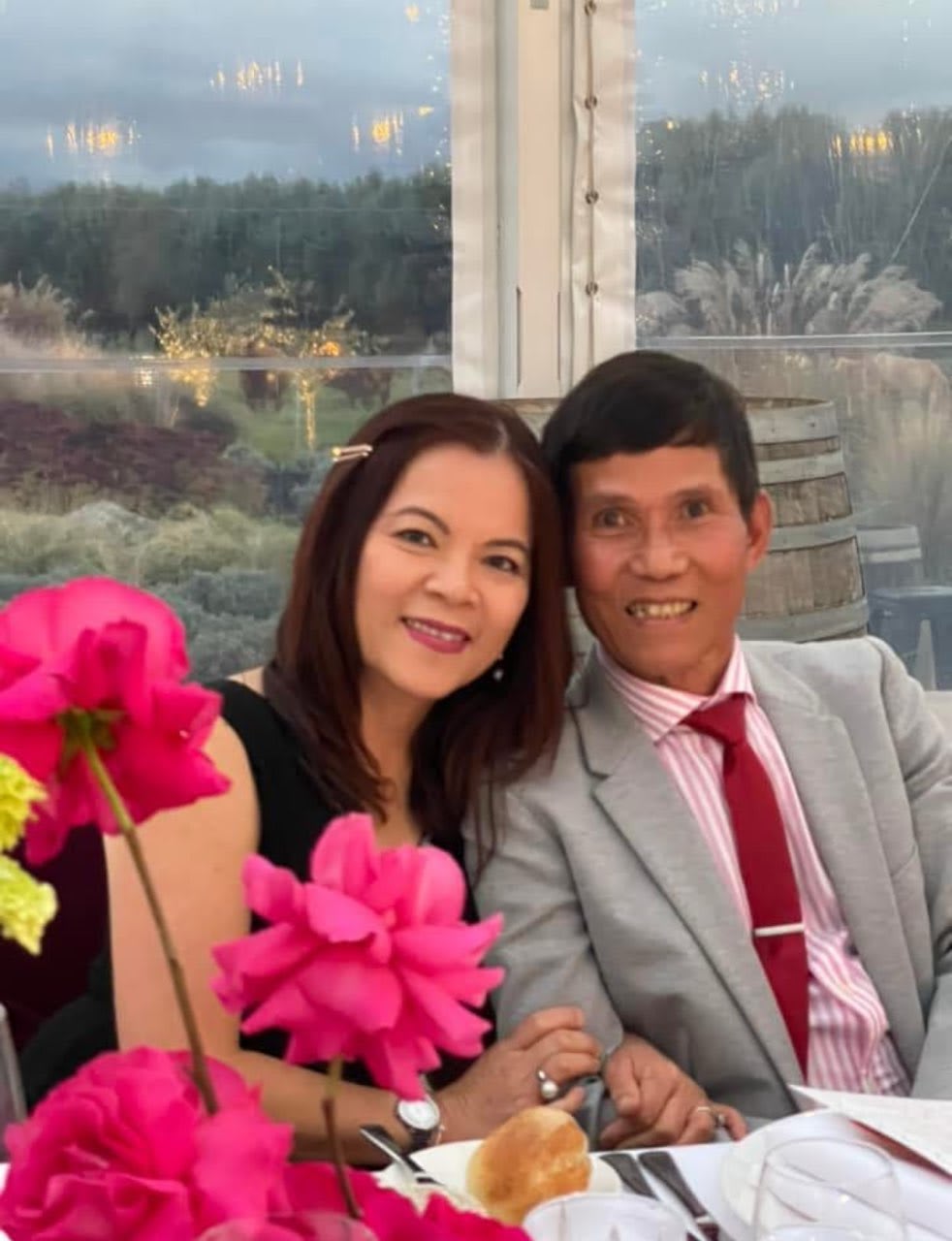 Couple smiling at dining table with pink flowers, man in suit, scenic outdoor view behind.