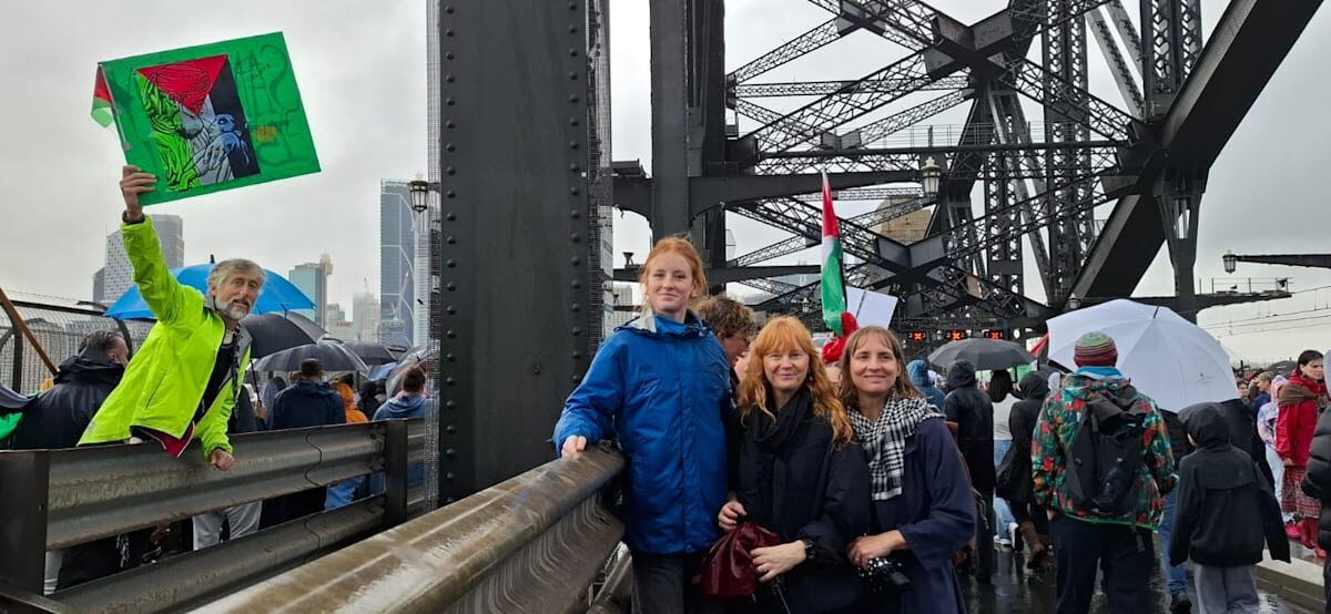 People Gathered On A Steel Bridge During A Cloudy Day Protest, Some Holding Signs And Umbrellas.