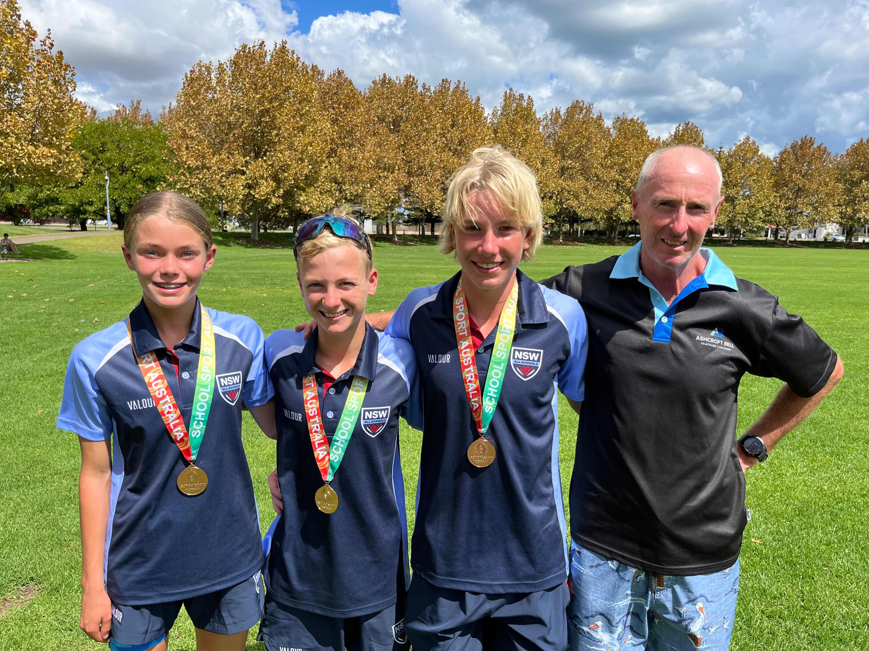Three Adolescents In Nsw Sports Uniforms With Gold Medals And An Adult Man On A Grassy Field.